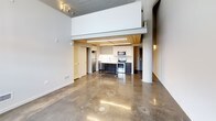This two-story space showcases an industrial-modern aesthetic characterized by polished concrete floors on the main level and a dramatic open layout with soaring ceilings. The ground floor centers around an open-plan living and kitchen area, where natural wood-planked ceilings warm the otherwise contemporary palette. The kitchen is well-appointed with stainless steel appliances, including a refrigerator, electric range, and over-the-range microwave, complemented by two-tone navy and white cabinetry, quartz countertops, and a central island. A structural column adds an authentic industrial character to the main living area, which also benefits from natural light through a wire-glass window. The upper level, accessed by a staircase, features carpeted flooring, a painted wood-plank ceiling, ceiling fan, and an arched architectural detail that adds visual interest. This floor includes a spacious walk-in closet with wire shelving and built-in lighting, as well as a clean, modern bathroom with a fully tiled walk-in shower, built-in shelf niche, and chrome fixtures. The overall ambiance is a cohesive blend of raw industrial elements and refined modern finishes throughout.