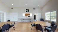 This single-story amenity facility is designed to serve as a versatile community hub, highlighted by two distinct functional areas. The centerpiece is a generously sized indoor pool room, distinguished by its soaring vaulted ceiling with exposed white structural beams and soft blue accent walls. Natural light streams in through a series of large arched and rectangular windows, creating a bright, airy atmosphere over the shimmering pool. An integrated hot tub is conveniently accessible at the pool's edge, bordered by brick tile detailing. Reclining lounge chairs line the perimeter, and potted plants add natural accents throughout the space.

The second key area is a multi-purpose gathering room featuring warm hardwood-style flooring, vaulted ceilings with recessed lighting, and an arched window that enhances the sense of openness. The room accommodates several round and rectangular tables with chairs, making it well-suited for events or meetings. A pass-through kitchenette with wood cabinetry, a refrigerator, and counter space supports catering and food service needs. Together, these spaces create a well-rounded facility ideal for recreational and social activities.