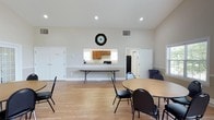 This single-story amenity facility is designed to serve as a versatile community hub, highlighted by two distinct functional areas. The centerpiece is a generously sized indoor pool room, distinguished by its soaring vaulted ceiling with exposed white structural beams and soft blue accent walls. Natural light streams in through a series of large arched and rectangular windows, creating a bright, airy atmosphere over the shimmering pool. An integrated hot tub is conveniently accessible at the pool's edge, bordered by brick tile detailing. Reclining lounge chairs line the perimeter, and potted plants add natural accents throughout the space.

The second key area is a multi-purpose gathering room featuring warm hardwood-style flooring, vaulted ceilings with recessed lighting, and an arched window that enhances the sense of openness. The room accommodates several round and rectangular tables with chairs, making it well-suited for events or meetings. A pass-through kitchenette with wood cabinetry, a refrigerator, and counter space supports catering and food service needs. Together, these spaces create a well-rounded facility ideal for recreational and social activities.