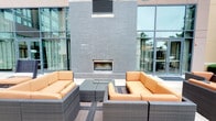 This contemporary single-floor space makes a strong architectural impression, blending indoor and outdoor living through thoughtful design. The standout feature is a striking outdoor patio lounge area anchored by a sleek, built-in linear fireplace set within a bold, dark textured brick wall. The fireplace serves as a dramatic focal point, drawing the eye and creating an inviting atmosphere for relaxation and social gathering. Flanking the fireplace on either side, large floor-to-ceiling windows and glass doors line the building's façade, allowing abundant natural light to flow through while maintaining a strong visual connection between the interior and exterior. The patio is furnished with modern wicker sectional sofas accented by warm orange cushions, arranged symmetrically around a low-profile coffee table, creating a cohesive and comfortable seating area. The neutral tones of the wicker and dark brick are enlivened by the vibrant upholstery, giving the space a welcoming, resort-like feel. The open-air layout, combined with the architectural details of the surrounding structure, lends the space a polished and modern ambiance well-suited for both casual lounging and social gatherings.