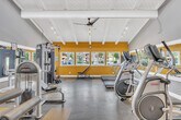 This state-of-the-art fitness center, located at 23251 Los Alisos Blvd in Lake Forest, CA, offers a compact yet comprehensive workout space within its 660 square feet. The facility's most striking feature is its vibrant yellow walls, which create an energizing atmosphere conducive to active pursuits. The space boasts impressively high ceilings with exposed white beams, enhancing the sense of openness and air circulation.

Large windows line one wall, flooding the area with natural light and providing views of the surrounding area. The flooring is a practical mix of carpet and hard surfaces, with yellow and gray square patterns adding visual interest and delineating different workout zones.

The gym is well-equipped with a variety of modern exercise machines, including treadmills, ellipticals, stationary bikes, and strength training equipment. Mirrors are strategically placed to assist with form and technique during workouts. The layout is thoughtfully designed to maximize the available space while ensuring ample room for movement between stations.

Attention to detail is evident in the lighting design, which combines natural light with recessed fixtures and a modern ceiling fan for comfort. The space also includes amenities such as a wall-mounted clock and informational signage, contributing to its functional and professional ambiance. Despite its single-floor layout, the gym's clever design and equipment selection provide a comprehensive fitness experience in a compact, efficient space.