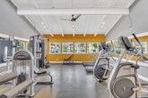 Located in Lake Forest, California, this fitness center offers a well-appointed workout space designed to support a comprehensive exercise routine. The room is defined by striking vaulted ceilings with exposed white-painted wooden beams, giving the space an open, airy feel that enhances the overall workout experience. A ceiling fan and track lighting run along the roofline, providing both ventilation and ample illumination throughout the day. Bold golden-yellow accent walls contrast with sage green lower sections, creating an energizing and motivating color palette. The flooring features a distinctive checkered pattern of gray speckled and yellow tiles, adding a dynamic visual element underfoot. Large windows along one wall flood the interior with natural light while offering views of the surrounding neighborhood. The gym is outfitted with a diverse array of cardio and strength-training equipment, including treadmills, elliptical machines, stationary bikes, and cable weight machines. Full-length mirrors along one wall visually expand the space and allow users to monitor their form during workouts. The fitness center is well-organized, making efficient use of its footprint to accommodate multiple users simultaneously, and is complemented by an adjacent entry corridor that provides a clean, welcoming transition into the space.