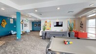This contemporary amenity lounge is thoughtfully designed to offer a dynamic and inviting social environment. The open-plan layout spans a generous single floor, creating a seamless flow between distinct activity and relaxation zones. At the forefront is a full-size pool table, serving as a central entertainment focal point and inviting friendly competition. Beyond it, a plush lounge area is arranged around a wall-mounted flat-screen television, with a large sectional sofa and accent chairs providing ample comfortable seating for gatherings or casual viewing. Patterned carpet throughout adds texture and warmth underfoot, complementing the clean white walls and coffered ceiling with recessed lighting. A striking teal-blue accent wall anchors a secondary seating nook, furnished with modern egg-style chairs and illuminated by stylish pendant lighting in aqua tones. Decorative wall art, sunburst mirrors, and tall potted plants are thoughtfully placed throughout, adding layers of personality and a lively aesthetic. The overall ambiance balances energetic social spaces with comfortable retreats, making this lounge a well-rounded and visually engaging communal destination.
