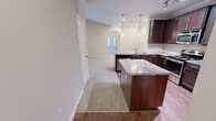 Located at 1084 W Oakland Ave in Johnson City, Tennessee, this single-story home offers a well-designed layout with functional living spaces throughout. The kitchen is a standout feature, showcasing rich dark wood cabinetry, granite countertops, a center island with additional storage, and pendant lighting overhead. The kitchen flows seamlessly into the living areas, which are finished with warm hardwood-style flooring that transitions to plush carpeting in the main living and bedroom spaces. Crown molding accents add a refined touch to the kitchen and adjoining areas. The living room is bright and inviting, featuring a ceiling fan, large windows with blinds, and a glass door providing direct access to the outdoors. The bedrooms are generously sized, each with carpeted floors, natural light from windows, and ample closet space. Two walk-in closets provide extensive storage, each equipped with wire shelving systems. The bathroom features tile flooring, a tub-shower combination with a curved curtain rod, a granite-topped vanity, and dark wood cabinetry consistent with the home's overall aesthetic. A dedicated laundry area houses a full-size washer with wire shelving above for added storage. The home's neutral color palette and cohesive finishes create a clean, comfortable living environment throughout.