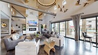 This single-story space masterfully combines rustic Western aesthetics with contemporary design elements across a generously sized, open floor plan. The architecture is defined by soaring vaulted ceilings supported by exposed steel beams, whitewashed stone accent walls, and polished concrete floors that flow seamlessly throughout the main areas.

The central great room serves as the heart of the space, anchored by a floor-to-ceiling stone fireplace wall with an inset electric fireplace and a mounted television above. Western-inspired artwork, sculptural bird wall installations, and antler-handled furnishings reinforce the thematic design. A sliding barn door adds a functional yet decorative touch.

Large sliding glass doors along one wall flood the interior with natural light and provide direct access to an outdoor terrace. Multiple seating arrangements accommodate both social gatherings and quieter, more intimate conversations. A secondary lounge area features floor-to-ceiling drapery, a tufted sectional, and warm wood accents for a cozier atmosphere.

The kitchen area includes a substantial island with a quartz countertop, tufted leather barstools, industrial pendant lighting with glass shades, and a gray cabinetry display hutch. A compact bistro nook and a dedicated workstation with a bookshelf-themed accent wall complete the space.