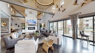 This single-story space masterfully combines rustic Western aesthetics with contemporary design elements across a generously sized, open floor plan. The architecture is defined by soaring vaulted ceilings supported by exposed steel beams, whitewashed stone accent walls, and polished concrete floors that flow seamlessly throughout the main areas.

The central great room serves as the heart of the space, anchored by a floor-to-ceiling stone fireplace wall with an inset electric fireplace and a mounted television above. Western-inspired artwork, sculptural bird wall installations, and antler-handled furnishings reinforce the thematic design. A sliding barn door adds a functional yet decorative touch.

Large sliding glass doors along one wall flood the interior with natural light and provide direct access to an outdoor terrace. Multiple seating arrangements accommodate both social gatherings and quieter, more intimate conversations. A secondary lounge area features floor-to-ceiling drapery, a tufted sectional, and warm wood accents for a cozier atmosphere.

The kitchen area includes a substantial island with a quartz countertop, tufted leather barstools, industrial pendant lighting with glass shades, and a gray cabinetry display hutch. A compact bistro nook and a dedicated workstation with a bookshelf-themed accent wall complete the space.
