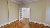 This single-floor space showcases a blend of classic architectural charm and timeless design. The room is finished with rich, warm hardwood floors that run throughout, adding a sense of warmth and continuity to the space. Crown molding frames the ceiling elegantly, lending a refined, traditional character to the interior.

A standout feature is the classic French door, complete with a multi-pane glass grid design, which opens into a dedicated closet area equipped with a built-in rod for storage. This detail adds both functionality and aesthetic appeal, elevating what might otherwise be a standard storage solution into an architectural highlight.

The walls are finished in a soft, neutral sage-green tone that creates a calm and inviting atmosphere, complementing the natural wood tones of the floor. Crisp white baseboards and door trim provide clean contrast and reinforce the traditional aesthetic throughout. The ceiling height feels generous, contributing to an open and airy ambiance.

Overall, the space combines practical storage solutions with thoughtful design details, offering a welcoming environment that balances classic style with everyday functionality. The cohesive use of neutral tones, natural materials, and traditional architectural elements makes this a versatile and well-appointed space.