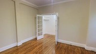 This single-floor space showcases a blend of classic architectural charm and timeless design. The room is finished with rich, warm hardwood floors that run throughout, adding a sense of warmth and continuity to the space. Crown molding frames the ceiling elegantly, lending a refined, traditional character to the interior.

A standout feature is the classic French door, complete with a multi-pane glass grid design, which opens into a dedicated closet area equipped with a built-in rod for storage. This detail adds both functionality and aesthetic appeal, elevating what might otherwise be a standard storage solution into an architectural highlight.

The walls are finished in a soft, neutral sage-green tone that creates a calm and inviting atmosphere, complementing the natural wood tones of the floor. Crisp white baseboards and door trim provide clean contrast and reinforce the traditional aesthetic throughout. The ceiling height feels generous, contributing to an open and airy ambiance.

Overall, the space combines practical storage solutions with thoughtful design details, offering a welcoming environment that balances classic style with everyday functionality. The cohesive use of neutral tones, natural materials, and traditional architectural elements makes this a versatile and well-appointed space.