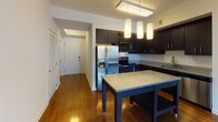 Located on the sixth floor at 4918 Saint Elmo Ave in the heart of Bethesda, MD, this single-level residence combines modern finishes with a functional, open layout. Rich hardwood flooring runs throughout the main living areas, reflecting natural light that pours in through floor-to-ceiling windows in the living room, which frame sweeping views of the surrounding Bethesda skyline.

The kitchen is a standout feature, outfitted with sleek dark cabinetry, granite countertops, and a full suite of stainless steel appliances including a French door refrigerator, electric range with microwave, and dishwasher. A freestanding island with a granite top and pendant lighting above provides additional prep space and a casual dining option, while a flush-mount ceiling fixture illuminates the full kitchen area.

The bedroom offers a comfortable retreat with plush carpet, clean neutral walls, and a wide horizontal window that captures urban views. Two bathrooms serve the unit — one features a glass-enclosed walk-in shower with white tile surround and brushed nickel fixtures, while the second includes a full soaking tub with tiled surround. Both bathrooms are finished with granite-topped vanities and neutral tile flooring. The entryway leads efficiently into the open living and kitchen space, making the layout feel cohesive and welcoming throughout.