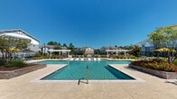 Located at 19216 Dr John Lambert Dr in Hammond, Louisiana, this outdoor amenity area serves as the centerpiece of a well-appointed residential community. The space is designed around an expansive, cross-shaped pool layout that includes multiple connected swimming pools with crystal-clear blue water, shallow sun shelves ideal for lounging, and ample deck space finished in light-toned pavers that reflect the Louisiana sunshine.

Surrounding the pools, a generous selection of chaise lounges and seating areas are thoughtfully arranged to maximize both relaxation and socializing. Colorful patio umbrellas in yellow and blue add a vibrant, resort-like atmosphere to the space. Raised planting beds featuring ornamental grasses and bold red flowering plants provide visual structure and color throughout the deck area.

A standout feature of the amenity is the covered outdoor cabana, constructed with exposed wood beams, a sloped roof, and brick accents. This structure houses an outdoor kitchen or bar area, offering shade and a gathering space for residents. White pergolas positioned along the pool perimeter provide additional shaded seating zones with a clean, classic aesthetic.

The entrance to the amenity is framed by lush landscaping, including mature trees and vibrant red flower beds, creating an inviting arrival experience. The multi-story residential buildings visible in the background complete the sense of a cohesive, well-designed community environment.