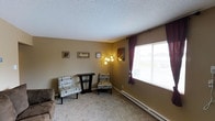 This single-floor unit features two bedrooms and one bathroom within a thoughtfully arranged floor plan. The living area is carpeted and bathed in natural light through a large window dressed with burgundy curtains and horizontal blinds, creating a warm and inviting atmosphere. Neutral tan walls provide a versatile backdrop, complemented by a textured popcorn ceiling. A baseboard heating unit runs along the exterior wall, ensuring efficient climate control. The space accommodates seating and accent furniture comfortably, with wall art adding a personal touch. The overall ambiance is cozy and understated, with practical finishes suited for everyday living. The single-floor layout provides easy accessibility throughout, connecting the living spaces without the need for stairs. Natural light plays a notable role in brightening the interior, particularly in the main living area where the oversized window draws in ample daylight. This unit presents a straightforward and functional living environment, well-suited for individuals or small households seeking a manageable, comfortable space.