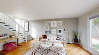 This two-storey home is thoughtfully designed with a cohesive neutral palette, warm parquet flooring throughout, and an open flow between its main living areas. The living room serves as the heart of the main floor, offering generous space for seating and entertaining. A large window paired with a glass door floods the room with natural light and provides direct access to an outdoor area, where greenery is visible through botanical-print curtains. A curated gallery wall anchors one side of the room, adding visual interest alongside the staircase leading to the upper level.

Adjacent to the living room, the dining area makes a bold design statement with a deep navy accent wall, a rattan sunburst mirror, and a pendant light overhead, creating an intimate yet striking atmosphere. A glimpse into the kitchen reveals white cabinetry and clean countertops.

Upstairs, the bedrooms continue the home's soft grey-green tones and parquet flooring. The primary bedroom features a window with outdoor views, a closet, and a decorative niche. A second bedroom offers a built-in closet and bright natural light. The bathroom is finished with light grey square tiles, a framed mirror, and clean white fixtures, completing the home's calm, curated aesthetic.