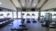 This fitness center combines rustic architectural charm with modern functionality. The vaulted ceiling is adorned with exposed wooden beams that add warmth and character to the space, while recessed lighting and a ceiling fan ensure comfort during workouts. Large floor-to-ceiling windows and glass doors line the walls, flooding the interior with natural light and offering open views of the surrounding outdoor area, creating an airy and motivating atmosphere.

The facility is outfitted with a solid selection of cardio equipment, including multiple treadmills and elliptical machines positioned to take full advantage of the exterior views. Free weights and a weight bench are available for strength training, rounding out the workout options. The dark, durable flooring provides a practical and cohesive foundation throughout the space.

Wall-mounted televisions are installed for entertainment during workouts. A mirrored wall on one side enhances the sense of spaciousness while serving a functional purpose for form-checking during exercise. Overall, the space strikes a balance between an inviting, rustic aesthetic and the practical amenities of a fully functional fitness facility.