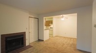 This single-floor residence offers a practical and comfortable layout designed for everyday living. The main living area is carpeted and centers around a brick-framed fireplace, adding a classic, cozy focal point to the room. An entry door positioned adjacent to the fireplace provides direct access to the space. The living area flows openly into a secondary room, also carpeted, which is outfitted with a ceiling fan and light fixture overhead, offering good air circulation and ambient lighting. A pass-through opening connects this area to the kitchen, which features warm wood-toned cabinetry, a white range with overhead ventilation, and a countertop that serves as a convenient transition between the cooking and living spaces. The kitchen's tile flooring contrasts with the carpet found throughout the rest of the unit, clearly delineating functional zones. The neutral wall tones and simple, clean finishes throughout create a straightforward and adaptable interior. Overall, the space presents a functional residential layout with traditional design elements, including the standout fireplace and open kitchen connection that lend both character and practicality to the home.
