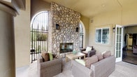 Located at 3875 Main in Chula Vista, this thoughtfully designed outdoor space blends Mediterranean-inspired architecture with comfortable, functional gathering areas. The centerpiece of the covered lounge is a striking floor-to-ceiling fireplace clad in natural river rock, flanked by arched iron-gated openings that frame views of the surrounding greenery. Warm stucco walls, soaring ceilings, and rounded architectural columns lend the space a refined yet inviting character. Wicker lounge seating arranged around the fireplace creates an intimate setting ideal for relaxed conversation or evening gatherings.

Adjacent to the covered lounge, a spacious open-air patio extends the entertaining area with multiple dining tables, shaded by large market umbrellas. A curved built-in bench wraps around a sleek rectangular gas fire pit, offering a dedicated social zone with panoramic views of the landscaped surroundings and open sky beyond. Textured aggregate concrete flooring ties the areas together, while lush drought-tolerant plantings and ornamental trees soften the perimeter. French doors connect the covered patio to an interior space, ensuring seamless indoor-outdoor flow. Together, these zones create a versatile and welcoming outdoor retreat suited for dining, lounging, and year-round entertaining.