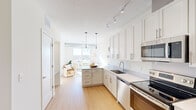 This well-appointed single-floor residence combines modern design with functional living. The open-concept kitchen is a standout feature, showcasing crisp white shaker cabinetry extending to the ceiling, light quartz countertops, a peninsula with pendant lighting, and a full suite of stainless steel appliances including a range, dishwasher, built-in microwave, and refrigerator. Track lighting overhead provides excellent task illumination throughout the kitchen. Light-toned hardwood flooring runs seamlessly across the entire unit, lending warmth and continuity to each space. The living area is generously proportioned and bathed in natural light, opening directly onto a private balcony through a full-height sliding glass door. The balcony offers expansive panoramic views of the surrounding cityscape, making it a compelling feature of the unit. Exposed industrial-style ductwork adds a subtle modern edge to the bedroom, which also benefits from large windows framing the same impressive city views. A spacious walk-in closet with wire shelving systems provides ample storage. Throughout the unit, neutral tones, clean lines, and an abundance of natural light create a bright, airy ambiance that is both functional and inviting.