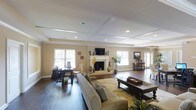 This single-floor space is defined by its standout coffered ceiling, which adds architectural depth and visual interest to the expansive main living area. Recessed lighting is thoughtfully integrated throughout the ceiling, providing even illumination across the room. Dark hardwood floors run throughout the space, offering a rich contrast against the warm, neutral-toned walls.

The focal point of the living area is a classic white-mantled fireplace, framed symmetrically and topped with a wall-mounted television, blending traditional design with modern functionality. A generously sized seating arrangement anchors the room, centered around a large coffee table atop a patterned area rug, creating a defined and comfortable lounge zone.

Multiple windows fitted with white blinds allow natural light to fill the space, enhancing its open and airy feel. A dedicated workspace with a desk and chair is positioned near the windows, offering a functional area within the living space. Additional accent furniture, including a console table and decorative greenery, contribute to the polished, well-appointed ambiance of this versatile and elegantly designed interior.