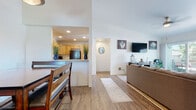 Located at 420 Activity Way in Oceanside, CA, this single-story coastal retreat exudes a relaxed beach ambiance throughout. The open-concept layout connects the living, dining, and kitchen areas seamlessly, all finished with warm wood-look flooring. The living room features vaulted ceilings, a ceiling fan, and sliding glass doors that open to a private balcony, allowing an abundance of natural light and outdoor access. The kitchen is outfitted with natural knotty wood cabinetry, light stone countertops, and black appliances, including a range and built-in microwave. A peninsula counter separates the kitchen from the dining area, which accommodates a wood dining table with bench seating beneath a pendant light. The home includes multiple bedrooms, each carpeted and decorated with coastal-inspired artwork. The primary bedroom offers sliding glass door access to the outdoors and a ceiling fan, while additional bedrooms feature large windows with views of the surrounding neighborhood and palm trees. Two bathrooms mirror each other in design, featuring knotty wood vanities, stone countertops, and tub/shower combinations — one styled with serene nautical accents and the other with a vibrant surf-themed curtain. The overall aesthetic is cohesive, light, and beach-inspired throughout.