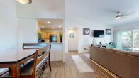 Located at 420 Activity Way in Oceanside, CA, this single-story coastal retreat exudes a relaxed beach ambiance throughout. The open-concept layout connects the living, dining, and kitchen areas seamlessly, all finished with warm wood-look flooring. The living room features vaulted ceilings, a ceiling fan, and sliding glass doors that open to a private balcony, allowing an abundance of natural light and outdoor access. The kitchen is outfitted with natural knotty wood cabinetry, light stone countertops, and black appliances, including a range and built-in microwave. A peninsula counter separates the kitchen from the dining area, which accommodates a wood dining table with bench seating beneath a pendant light. The home includes multiple bedrooms, each carpeted and decorated with coastal-inspired artwork. The primary bedroom offers sliding glass door access to the outdoors and a ceiling fan, while additional bedrooms feature large windows with views of the surrounding neighborhood and palm trees. Two bathrooms mirror each other in design, featuring knotty wood vanities, stone countertops, and tub/shower combinations — one styled with serene nautical accents and the other with a vibrant surf-themed curtain. The overall aesthetic is cohesive, light, and beach-inspired throughout.