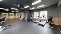 This fitness center offers a comprehensive, single-floor workout facility designed to accommodate a wide range of exercise needs. The main workout area features durable rubber flooring throughout, providing a safe and functional surface for both strength training and cardio activities. The space is finished with two-tone walls — a muted blue-gray upper half and a warm beige lower half — complemented by a dark ceiling that gives the facility a modern, focused atmosphere. Flush-mounted fluorescent lighting and ceiling fans ensure the space remains bright and well-ventilated during workouts. A dedicated strength training zone is lined with full-length mirrors and houses multiple cable machines, weight benches, and resistance equipment, allowing users to train various muscle groups. On the opposing side, a row of treadmills and elliptical machines is positioned along large windows that frame views of the outdoors, filling the area with natural light. Additional open floor space provides room for stretching, mat work, and group fitness accessories such as step platforms. The facility also includes adjacent tiled rooms accessible through open archways, offering supplemental areas for changing or additional activities. An exit door provides convenient access to the exterior.