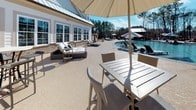 This single-story outdoor amenity space is designed to deliver a resort-like experience, combining a large swimming pool with generous surrounding hardscape finished in a textured, neutral-toned concrete coating. The pool area is well-appointed with multiple lounge chairs and shade umbrellas positioned along the pool deck, offering ample space for relaxation. A backdrop of tall pine trees provides a natural screen and a sense of privacy.

The covered patio area is architecturally detailed with exposed wood-beam ceilings, white columns, and a combination of brick and board-and-batten siding that lends a refined yet approachable aesthetic. This shaded zone includes a bar counter with high stools, creating a functional social gathering space. High-top bistro tables with wicker chairs are also distributed throughout, accommodating both casual conversation and outdoor dining.

Linear drainage channels are integrated cleanly into the deck, reflecting thoughtful infrastructure design. Large windows on the adjacent building allow natural light to flow into the interior while maintaining visual connectivity to the outdoor space. The overall ambiance is bright, open, and well-suited for community gatherings or leisure throughout the day.