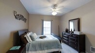 This bedroom offers a well-appointed and comfortable retreat, designed with both function and style in mind. The room is finished with soft wall-to-wall carpeting underfoot, contributing to a warm and inviting ambiance throughout. Neutral-toned walls provide a calm backdrop that complements the rich, dark wood furniture, including a sleigh bed frame and a spacious dresser with a matching mirror. The dresser provides generous storage and display space, with decorative accents adding a personal touch. A ceiling fan with an integrated light fixture is centrally positioned, ensuring effective air circulation and ambient lighting. Natural light filters into the room through a window dressed with light blue curtains, offering a bright and airy feel while maintaining privacy. A decorative wall piece adds a subtle artistic element to the space. The overall layout is thoughtfully arranged to maximize usability, with ample space around the bed for comfortable movement. The combination of soft textiles, coordinated furnishings, and natural light creates a restful and aesthetically cohesive environment well-suited for relaxation.