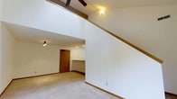 This two-story space is defined by its dramatic vaulted ceilings, which lend a sense of openness and volume to the main living area. Exposed wooden beams accent the upper ceiling, adding a warm, structural detail that contrasts with the clean white walls throughout. A ceiling fan is positioned at the upper level, providing both function and a casual aesthetic touch.

The main level features wall-to-wall carpeting in a neutral tone, contributing to a soft, comfortable feel underfoot. Warm wood trim runs along the baseboards and staircase railing, providing cohesion and a classic finish. Recessed lighting and a small multi-bulb fixture illuminate the lower level, ensuring adequate lighting throughout the space.

A staircase with a wood-trimmed railing leads to the upper level, which opens to an overlook or loft-style area visible from below. The entry area features a solid wood door with standard hardware, framed by white walls. A built-in half-wall or counter detail near the entry adds an architectural element that subtly defines the transition between spaces. Overall, the layout is open and functional, with a neutral palette suited for a variety of uses.