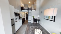 Located at 6105 Roosevelt Way NE in Seattle's vibrant Roosevelt neighborhood, this thoughtfully designed loft-style unit makes exceptional use of vertical space. Soaring ceilings define the main living area, drawing the eye upward to a mezzanine sleeping loft accessible via a sleek black metal ladder with matching railing — a striking architectural focal point that adds both function and character to the space.

The open-plan layout seamlessly integrates the kitchen, living, and work areas. The compact kitchen is efficiently outfitted with stainless steel appliances, including a microwave and refrigerator, complemented by clean white cabinetry and a dark-toned dresser unit that adds contrast and storage. The living area features warm hardwood-style flooring throughout, anchored by a patterned area rug that adds texture and depth.

A dedicated workspace with a desk and chair sits adjacent to the living area, making the unit well-suited for remote work or study. The neutral grey and white color palette, accented by black fixtures and hardware, gives the space a contemporary, cohesive aesthetic. Natural and ambient lighting work together to keep the interior feeling bright and open. This unit offers a smart, modern living experience in one of Seattle's most walkable and connected neighborhoods.
