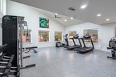This single-level fitness center in Madison, Alabama offers a spacious and well-appointed workout environment designed to support a full range of exercise routines. The defining architectural feature is the dramatic vaulted ceiling with exposed dark wood beams that span the length of the room, lending the space a sense of openness and volume. Large modern ceiling fans hang from the pitched roofline, ensuring consistent air circulation throughout. The flooring is a distinctive combination of tiled sections in cool blue and neutral gray tones, complemented by warm hardwood in the entry and peripheral zones, creating a visually dynamic layout. Bold navy blue accent walls provide a striking contrast to the crisp white walls, adding a contemporary energy to the atmosphere. The fitness floor is comprehensively equipped with treadmills, ellipticals, stationary and spin bikes, cable machines, a power rack, free weights, a dumbbell rack, and a punching bag, catering to both cardio and strength training needs. Mounted televisions are positioned throughout for entertainment during workouts. Large windows along the walls allow ample natural light to fill the space. A full-length wall mirror expands the visual depth of the room while supporting form-focused training. The overall ambiance is modern, motivating, and meticulously maintained.
