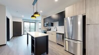 This contemporary single-floor unit showcases a thoughtfully designed open-concept layout with clean lines and modern finishes throughout. The kitchen is a standout feature, equipped with a full suite of stainless steel appliances including a French door refrigerator, electric range, built-in microwave, and dishwasher. Two-tone cabinetry pairs matte dark upper cabinets with light wood lower cabinets, complemented by a classic white subway tile backsplash and a central island with a white countertop and pendant lighting above. The living area is bright and open, featuring light wood-style plank flooring, exposed metallic ductwork as an industrial accent, and large sliding glass doors that lead to a private balcony with views of the surrounding building community. Bedrooms offer plush carpet flooring and large windows that bring in natural light, with one bedroom featuring a full-height picture window overlooking the exterior. The bathroom is finished with a wide light wood vanity, white countertop, black-framed rectangular mirror, and modern black hardware. Dark trim accents tie together the overall aesthetic, creating a cohesive, urban-modern ambiance throughout the space.
