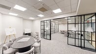 This thoughtfully designed office suite spans a single floor and comprises five functional rooms, each defined by a cohesive modern aesthetic. The standout architectural feature is the bold black-framed glass partition system that divides the space, creating distinct zones while maintaining visual connectivity and an open, airy feel throughout.

The primary conference room is anchored by a sleek oval dark table surrounded by contemporary upholstered chairs, a wall-mounted flat-screen display, and presentation easels — making it well-suited for meetings and collaborative sessions. An adjacent larger room features a long natural wood communal table with ample seating, ideal for team gatherings or working sessions.

Several smaller enclosed rooms, each accessed through the glass partition doors, provide more intimate settings furnished with compact tables and chairs, suitable for focused work or one-on-one meetings. Colorful artwork accents select rooms, adding visual interest to the otherwise clean, neutral palette.

Throughout the suite, light-toned patterned carpet, crisp white walls, and drop-tile ceilings with recessed panel lighting create a consistently bright and professional atmosphere. The interconnected layout, unified material choices, and transparent partitions foster a sense of openness and collaboration while still offering privacy where needed.