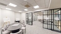 This thoughtfully designed office suite spans a single floor and comprises five functional rooms, each defined by a cohesive modern aesthetic. The standout architectural feature is the bold black-framed glass partition system that divides the space, creating distinct zones while maintaining visual connectivity and an open, airy feel throughout.

The primary conference room is anchored by a sleek oval dark table surrounded by contemporary upholstered chairs, a wall-mounted flat-screen display, and presentation easels — making it well-suited for meetings and collaborative sessions. An adjacent larger room features a long natural wood communal table with ample seating, ideal for team gatherings or working sessions.

Several smaller enclosed rooms, each accessed through the glass partition doors, provide more intimate settings furnished with compact tables and chairs, suitable for focused work or one-on-one meetings. Colorful artwork accents select rooms, adding visual interest to the otherwise clean, neutral palette.

Throughout the suite, light-toned patterned carpet, crisp white walls, and drop-tile ceilings with recessed panel lighting create a consistently bright and professional atmosphere. The interconnected layout, unified material choices, and transparent partitions foster a sense of openness and collaboration while still offering privacy where needed.