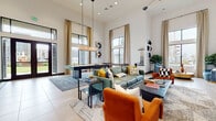 This thoughtfully designed single-story amenity space combines bold contemporary styling with generous proportions throughout. The main lobby welcomes visitors with dramatic soaring ceilings, floor-to-ceiling glass entry doors, and an elegant tiered chandelier that anchors the space. A built-in service counter along one wall offers a functional reception area, while plush teal velvet lounge chairs and a geometric-patterned area rug create an inviting seating arrangement.

Adjacent to the lobby, a spacious lounge and recreation area features equally impressive ceiling heights, flooded with natural light through oversized black-framed windows dressed with floor-length gold curtains. A billiards table positioned at the center of this room serves as a social focal point, complemented by a sleek linear pendant light overhead. Comfortable seating — including a sage green leather sofa, vibrant burnt orange armchairs, and eclectic accent pieces — gives the space a curated, magazine-worthy aesthetic. Neutral large-format tile flooring runs throughout.

A dedicated café-style nook features ornate patterned encaustic tile flooring, intimate round tables with velvet chairs, and access to a coffee station — offering a quieter retreat with courtyard views through expansive windows.