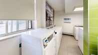 This 400 square foot laundry facility, located at 10 Roanoke Rd in Toronto, is a well-designed, single-floor space that prioritizes functionality and efficiency. The room features a clean, modern aesthetic with white walls, bright lighting, and large-format tile flooring. Multiple washing machines and dryers line the walls, providing ample capacity for residents' laundry needs. A central island with a white countertop offers a convenient folding and sorting area. The space incorporates both wooden and white cabinetry for storage and organizational purposes. Safety features include a prominently displayed fire extinguisher and clear signage. A mix of white, gray, and vibrant green accent walls adds visual interest while maintaining a professional atmosphere. Informative posters and bulletin boards are strategically placed to communicate important information to users. The facility also includes a small seating area and bookshelves, creating a multi-functional space for residents to utilize while doing laundry. Large windows allow natural light to enter, enhancing the overall brightness of the room. This thoughtfully designed laundry facility combines practicality with comfort, serving as an essential amenity for the residential community.