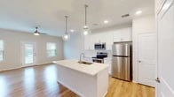 This two-floor home is designed with a clean, contemporary aesthetic that prioritizes bright, open living. The main level centers around an open-concept kitchen and living area finished with light hardwood flooring that extends throughout. The kitchen is well-appointed with white shaker-style cabinetry, a white quartz countertop, subway tile backsplash, and a full suite of stainless steel appliances including a refrigerator, electric range, and over-the-range microwave. A central island with an integrated sink provides additional workspace and seating potential, while two globe pendant lights hang above, adding a stylish focal point. Recessed lighting and a ceiling fan in the living area ensure the space remains bright and comfortable. Multiple windows allow natural light to filter in, creating an airy, welcoming atmosphere. The upper level features carpeted bedrooms with neutral-toned walls, including a primary bedroom with access to a spacious walk-in closet outfitted with wire shelving systems. The primary bathroom offers a double-sink vanity with white cabinetry, quartz countertops, and a full-width mirror. A secondary bathroom includes a tub-shower combination with white tile surround and hardwood-style flooring, maintaining the home's consistent, polished finish throughout.