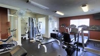 This single-floor fitness center offers a comprehensive workout environment designed to accommodate a range of exercise needs. The space features a practical and organized layout, with dedicated areas for both strength training and cardiovascular equipment. Strength training options include a multi-station cable machine and a weight bench, while the cardio section is equipped with treadmills, an elliptical, and a recumbent bike, providing ample variety for users of all fitness levels. The flooring throughout is dark rubber, offering durability and safety during workouts. Walls are painted in a combination of warm beige and a bold deep red accent, lending the space an energizing atmosphere. Ceiling-mounted fluorescent lighting ensures the room is well-lit, and wall-mounted fans contribute to ventilation and comfort during use. A large mirror on one wall enhances the sense of space and allows users to monitor their form. Natural light filters in through windows, adding brightness to the environment. The entrance features a glass-panel door, providing easy access. The textured ceiling and clean, functional design make this fitness center a practical and inviting facility for daily exercise.
