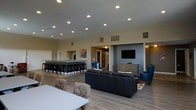 This single-floor space is designed as an open-concept common area that seamlessly blends multiple functional zones into one cohesive environment. The layout encompasses a generous lounge area anchored by a sleek black leather sofa and complemented by striking blue accent chairs, creating a welcoming gathering spot. A wall-mounted flat-screen television and a wood-toned media console serve as a focal point for the media area, making it ideal for entertainment and social gatherings.

Adjacent to the lounge, a bar-height counter with multiple bar stools provides additional seating and a casual dining or socializing option. Large mirrors behind the bar area enhance the sense of space and reflect the warm ambient lighting throughout. A white dining table with upholstered seating occupies the foreground, offering a dedicated area for meals or meetings.

The space is finished with wood-style luxury vinyl flooring that runs throughout, lending warmth and continuity to the open floor plan. Recessed ceiling lighting provides bright, even illumination across the entire area, while the neutral beige and gray wall tones create a clean, contemporary backdrop. Accent artwork adds a touch of color and personality. Overall, the space presents a versatile, well-appointed environment suited for community use and social interaction.