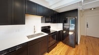 Studio S2 is a thoughtfully designed studio space that maximizes functionality within a compact, single-floor footprint. The kitchen is a standout feature, showcasing bold dark cabinetry paired with crisp white countertops and a classic white tile backsplash. Black appliances — including an electric range, over-the-range microwave, dishwasher, and full-size refrigerator — create a cohesive, modern aesthetic, while a gold-toned gooseneck faucet adds a refined accent. Warm hardwood flooring flows throughout the main living area, enhancing the open and airy feel of the space. Exposed structural elements on the ceiling lend an industrial character that complements the clean, contemporary design. The bathroom continues the bold black-and-white design language, featuring a dark vanity with white countertop, a round gold-framed mirror, and a glass-enclosed walk-in shower with black framing and gold fixtures. White tile flooring with subtle mosaic detail adds texture underfoot. The main living area provides flexible open space with good natural light filtering in, along with access to a closet and the bathroom, keeping the layout efficient and user-friendly.
