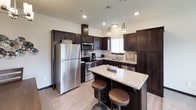 This two-story residence is thoughtfully designed with a functional and modern aesthetic throughout. The kitchen serves as a central highlight, featuring rich dark-stained cabinetry, granite-style countertops, and a full suite of stainless steel appliances including a refrigerator, electric range, and built-in microwave. A center island with pendant lighting and bar seating provides additional workspace and a casual dining option, while light wood-look flooring ties the space together.

The living area offers a comfortable, open feel with carpet flooring, a large window overlooking a green outdoor landscape, and direct access to the front entry. Multiple bedrooms are present across both floors, each carpeted and designed for restful use, with windows providing natural light.

Bathrooms include a full bath with a tub-shower combination, granite vanity, hexagonal tile flooring, and an adjacent walk-in closet with wire shelving. A powder room with a pedestal sink offers added convenience. Dark trim and hardware create consistent contrast throughout the space.

In-unit laundry is included, with a side-by-side washer and dryer complemented by overhead shelving for storage. The overall design balances practicality with clean, contemporary finishes.