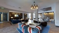 This expansive single-floor lounge is designed to accommodate a variety of social activities across several distinct zones. At its center, a bold Union Jack-upholstered tufted sofa anchors the main seating area, paired with a glass-top coffee table and dark leather club chairs, creating an eye-catching focal point. A rustic chandelier with cylindrical candle-style bulbs hangs overhead, adding warmth to the bright, open layout.

Built-in wooden shelving lines one wall, framing a wall-mounted flat-screen TV and providing a cozy, library-like atmosphere for relaxed gatherings. Large windows throughout allow abundant natural light to fill the space, complementing the light-toned walls and hardwood flooring.

Deeper into the room, a billiards table and additional high-top seating define a game area, while a textured accent wall adds dimension and visual interest. Recessed lighting throughout ensures consistent illumination across all zones. The overall ambiance balances a sophisticated, contemporary aesthetic with casual, inviting comfort, making it well-suited for communal use.