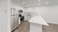This single-floor unit showcases a clean, contemporary design defined by a bright white color palette and warm wood-look flooring that runs consistently throughout the space. The open-concept kitchen serves as a central highlight, featuring flat-front white cabinetry extending to the ceiling for ample storage, a white quartz peninsula countertop ideal for casual dining or food preparation, and a full suite of stainless steel appliances including a refrigerator, electric range, built-in microwave, and an undermount sink. Recessed lighting illuminates the kitchen and living area, enhancing the modern aesthetic.

The bedroom is generously proportioned, finished with the same dark wood-look flooring and crisp white walls. A ceiling fan with integrated light fixture provides both comfort and illumination. Two doorways lead from the bedroom — one opening to the main living area and another providing direct access to a dedicated walk-in closet, offering notable storage convenience.

The bathroom maintains the unit's clean design language with white subway tile lining the tub surround, dual built-in niche shelves for storage, chrome fixtures including a square rainfall showerhead, and dark tile flooring that grounds the otherwise all-white space. Overall, the unit presents a cohesive, move-in-ready aesthetic throughout.