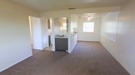 This single-floor space is designed with an open-concept layout that connects the main living area to the kitchen, creating a cohesive and functional environment. The living area is finished with wall-to-wall carpeting in a neutral tone, contributing to a comfortable and understated atmosphere. A ceiling fan is centrally positioned overhead, providing both ventilation and a modest lighting fixture.

The kitchen is defined by a half-wall peninsula that separates it from the living space while maintaining visual openness between the two areas. The kitchen features dark cabinetry with a light countertop surface, and natural light filters in through a window, brightening the workspace. A mirror or reflective surface near the kitchen adds depth to the area.

Adjacent to the kitchen, a utility or bathroom space is partially visible, equipped with additional cabinetry and a sink, suggesting a functional wet area nearby. Louvered closet doors are visible along one wall, offering built-in storage. The overall color palette is light and neutral throughout, with white walls that enhance the sense of openness. The layout prioritizes practicality, with clearly defined zones flowing naturally from one to the next.