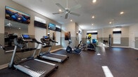 Located at 3995 S. Hamilton Rd. in Groveport, Ohio, this fitness center offers a thoughtfully designed, single-floor workout facility suited for a wide range of exercise routines. The space features a durable rubber flooring surface throughout, providing both safety and comfort during workouts. Neutral two-tone walls with a gray wainscoting detail give the interior a clean, modern aesthetic.

The cardio zone is well-appointed with multiple treadmills and stationary bikes, each equipped with individual digital displays for performance tracking. Wall-mounted flat-screen televisions are positioned throughout the space, ensuring an engaging workout experience. Ceiling fans complement the recessed lighting, promoting air circulation and a comfortable environment.

Full-length mirrors are strategically placed along the walls, enhancing the sense of openness and allowing users to monitor their form. The free weights section includes a complete dumbbell rack paired with an adjustable workout bench, catering to strength training needs. Additional functional training equipment includes exercise balls, resistance tools, and a cable machine.

Natural light filters through multiple windows fitted with blinds, contributing to a bright and airy atmosphere. A dedicated entry door with exterior views adds to the welcoming feel of the space. The overall layout is open and spacious, allowing for comfortable movement between equipment zones.