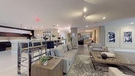 This single-floor amenity space is designed with a contemporary aesthetic, offering a series of well-defined zones that collectively create a versatile and social environment. The open layout flows seamlessly from one area to the next, beginning with a spacious lounge area furnished with plush gray sofas, upholstered accent chairs, and wood-finished coffee tables arranged atop textured area rugs. Recessed and track lighting illuminate the neutral-toned walls, contributing to a warm yet polished ambiance.

A high-top bar counter with sleek metal barstools serves as a central gathering point, connecting the lounge to the kitchen area. The kitchen features a striking two-tone design with dark lower cabinetry and crisp white upper cabinets, complemented by a clean white countertop island, pendant lighting, large-format polished tile flooring, and stainless steel appliances including a refrigerator and microwave.

Further into the space, a dedicated lounge nook showcases wood-look tile flooring, a light gray sofa, and Barcelona-style chairs beneath a large decorative compass artwork. Adjacent to this is a game room equipped with a ping-pong table, foosball table, a wall-mounted flat-screen TV, and black-and-white artwork, making it an engaging recreational retreat.