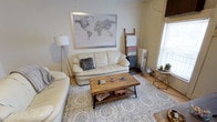 This single-floor space offers a comfortable and thoughtfully styled living area that blends vintage charm with casual, eclectic décor. The living room serves as the heart of the space, centered around a cream leather sofa and loveseat ensemble arranged atop a patterned area rug with intricate medallion detailing. A rustic wood-and-metal coffee table anchors the seating area, adding an industrial touch that contrasts warmly with the softer furnishings. A large framed world map print adorns the main wall, lending a sense of wanderlust and personality to the room. Natural light filters in through a window fitted with blinds, illuminating the space alongside a classic tripod floor lamp. A decorative leaning ladder holding neatly draped throw blankets and towels adds both function and style, while a traditional cast-iron radiator nearby speaks to the building's classic architectural character. A small gold geometric side table and potted greenery complete the corner, adding a modern accent. The light-toned walls and hardwood-style flooring contribute to an open, airy atmosphere throughout the living area, making it an inviting and functional space for everyday living.
