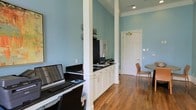 This single-floor space combines practical functionality with classic design elements. The interior is characterized by soft blue walls that create a calm, professional atmosphere, complemented by crisp white crown molding and white-painted architectural columns that add a traditional touch. Warm hardwood flooring runs throughout, lending a sense of continuity and comfort.

A dedicated workstation area is positioned along one wall, featuring a dark countertop surface housing computer equipment, including a laptop and keyboard setup. Adjacent to this, built-in white cabinetry with recessed panel doors and integrated drawer pulls provides ample storage, extending along the lower portion of the wall for an organized, streamlined appearance.

A structural column anchors the workspace, subtly dividing the area without fully closing off sight lines. Further into the space, a seating area is visible, with a round table and chair positioned near a paneled white door, suggesting the area can serve multiple functions. Recessed lighting in the ceiling provides even illumination throughout, enhancing the clean, well-maintained character of the overall environment.