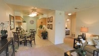 This single-floor residence offers a thoughtfully designed open-concept layout that seamlessly connects the living, dining, and kitchen areas, creating a cohesive and functional living environment. The living and dining spaces are anchored by a ceiling fan with light fixture, providing both comfort and ambient lighting. Soft, neutral-toned carpet flows throughout the main living areas, contributing to a warm and inviting atmosphere.

The dining area comfortably accommodates a full table and chair set, ideal for everyday meals or entertaining. Adjacent to the dining space, the kitchen features warm wood-toned cabinetry, light countertops, and standard appliances including a refrigerator. The kitchen is finished with tile or hard flooring, offering a practical contrast to the carpeted living zones.

A central partial-wall column defines the transition between the open living area and the hallway, which leads to the bedroom. The bedroom, visible through the corridor, is generously proportioned and carpeted, with its own ceiling fan and ample room for furnishings. The textured ceiling finish is consistent throughout, adding subtle architectural character. Overall, the space presents a comfortable, functional, and well-connected floor plan suited to everyday residential living.