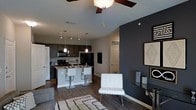This single-floor space showcases an open-concept design that seamlessly connects the kitchen, dining, and living areas into one cohesive living environment. The kitchen is well-appointed with dark cabinetry, stainless steel appliances including a gas range and refrigerator, and a central island with a light countertop that doubles as a breakfast bar with white bar seating. Pendant lighting hangs above the island, adding focused illumination and a modern aesthetic touch.

The living area features a ceiling fan with integrated lighting, contributing to both comfort and ambiance. A bold dark accent wall anchors the living space, creating a striking contrast against the lighter walls and ceilings. Rich, dark hardwood-style flooring runs throughout, lending warmth and continuity across the open floor plan. A textured area rug and a sleek white accent chair add layers of style to the space.

The layout offers a strong sense of flow and functionality, with a doorway visible in the background suggesting additional rooms. Overall, the space presents a contemporary, well-designed interior with thoughtful details and a balanced mix of light and dark tones.