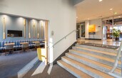 This single-floor common area showcases a thoughtfully designed, nautical-inspired aesthetic that sets a welcoming tone throughout. The entry foyer features a grand staircase with stone-finished steps and a sleek metal handrail, flanked by an elevator for convenient accessibility. Soaring ceilings with exposed dark beams and recessed lighting create an open, airy atmosphere across the entire space.

The lounge areas are divided into distinct yet connected zones. One seating nook features bold yellow upholstered chairs arranged around a round marble-top table, bathed in natural light from expansive floor-to-ceiling windows. An adjacent lounge offers a plush sectional sofa with a glass coffee table, framed coastal artwork, and generous window views of the surrounding neighborhood.

A dedicated café-style seating area incorporates striped blue banquette seating, live-edge wood tables, and decorative golden oars mounted on a blue accent wall — reinforcing the nautical theme with texture and character. A wall-mounted flat-screen TV and additional lounge chairs with woven details complete the communal gathering space. Blue-toned carpeting ties each area together, complementing the coastal color palette throughout.