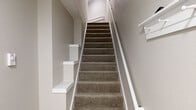 This two-storey residence is thoughtfully designed with a clean, contemporary aesthetic that carries consistently throughout both levels. The main floor features warm wood-look laminate flooring that runs through the open-concept living and kitchen areas, creating a cohesive and inviting atmosphere. Recessed lighting illuminates the space efficiently, complementing the neutral beige and white tones found on the walls and trim.

The kitchen stands out as a focal point on the main level, outfitted with full-height white shaker-style cabinetry, stainless steel appliances including a refrigerator, electric range, built-in microwave, and dishwasher. A large white quartz island with an overhang provides additional workspace and casual seating potential. A deep undermount sink with a matte black faucet adds a modern contrast. The living area beyond the kitchen includes a fireplace, adding warmth and character to the space.

A staircase with a white-painted newel post and handrail connects the two levels, transitioning from the wood-look flooring below to plush grey-brown carpet above. The upper level features carpeted rooms finished in soft neutral tones, with windows dressed in vertical blinds allowing natural light. Baseboard heating is present in the upper-level rooms, ensuring year-round comfort throughout the home.